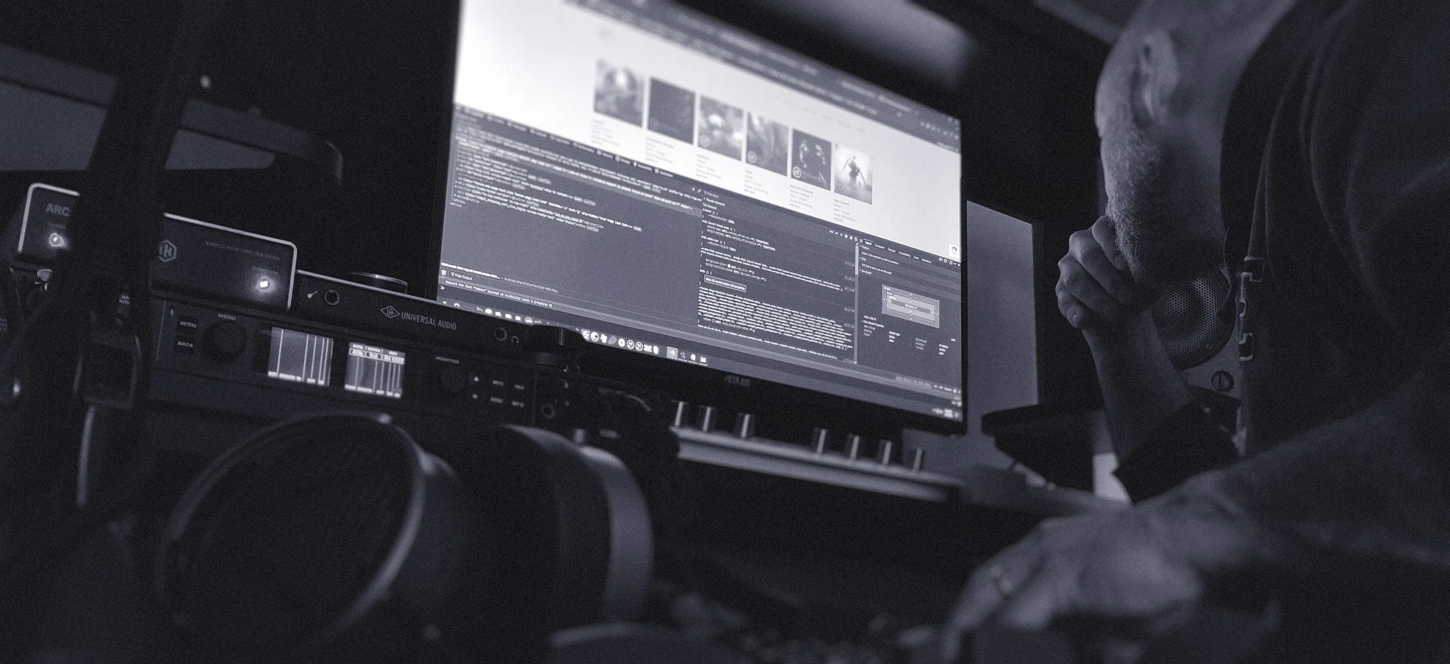 A person leans over a desk, surrounded by audio equipment, looking at a large computer monitor displaying code and images in a dimly lit workspace, capturing a moment of creative development for music artists.