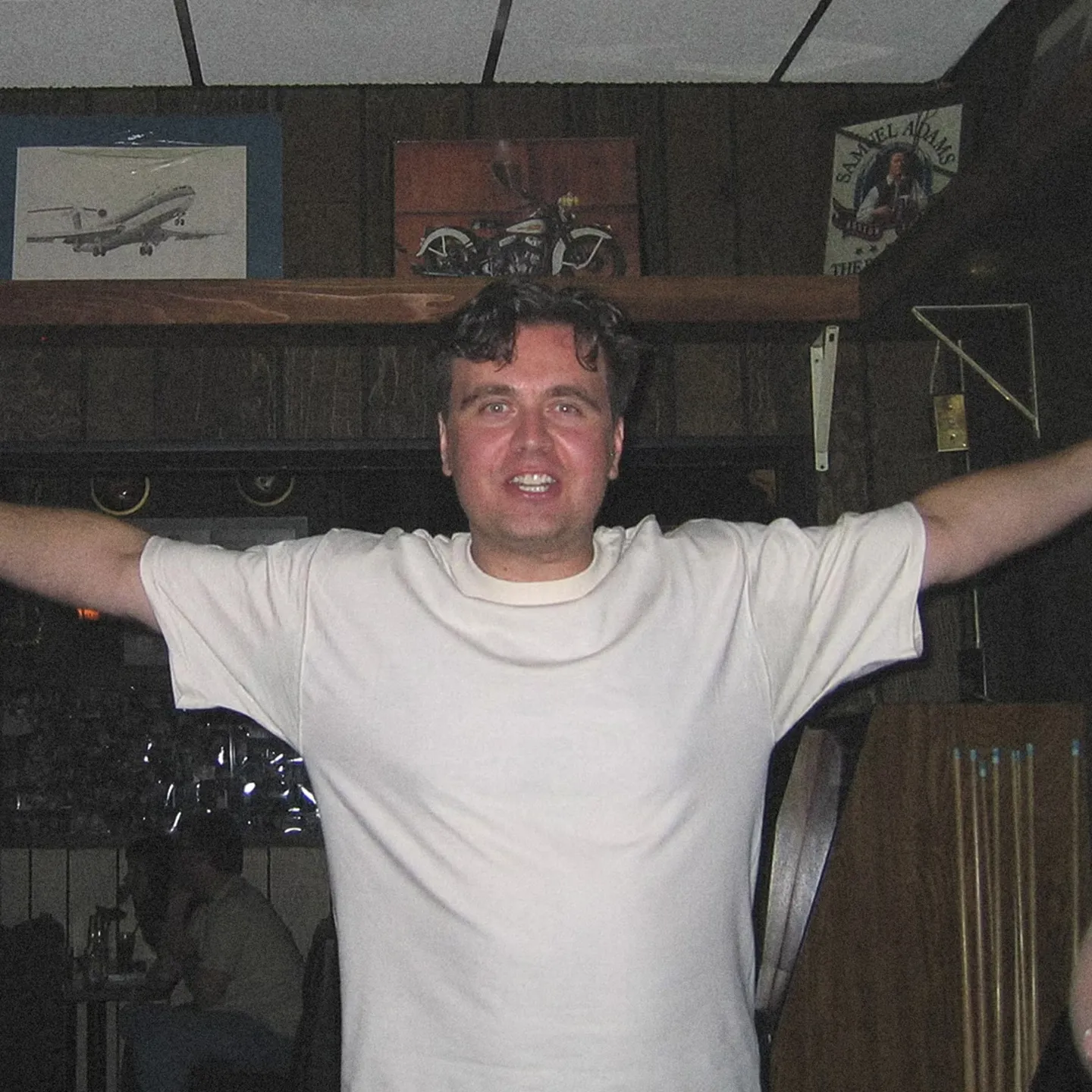 A smiling man stands in a dimly lit bar with his arms open wide. He wears a white shirt, exuding a welcoming and cheerful vibe.